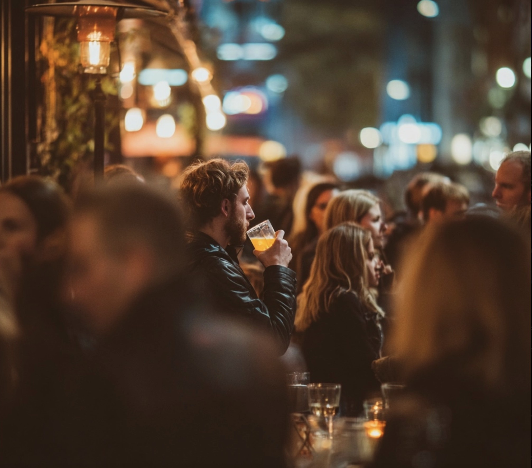 Rot days - A man with blonde hair and a leather jacket taking a sip of a drink at a crowded outdoor patio bar at night, surrounded by blurred figures and warm bokeh city lights.