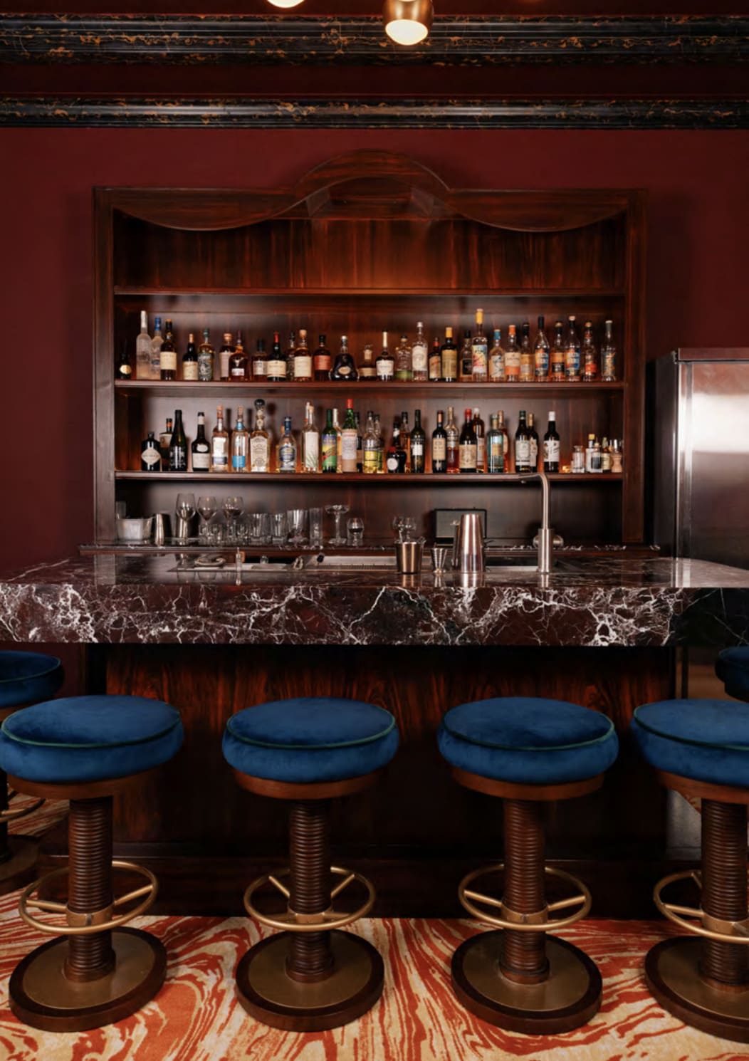 Vue intérieure du bar à cocktails Bar des Clos, speakeasy intimiste au décor rouge cerise, tabourets velours bleu et comptoir en marbre sombre à Paris.